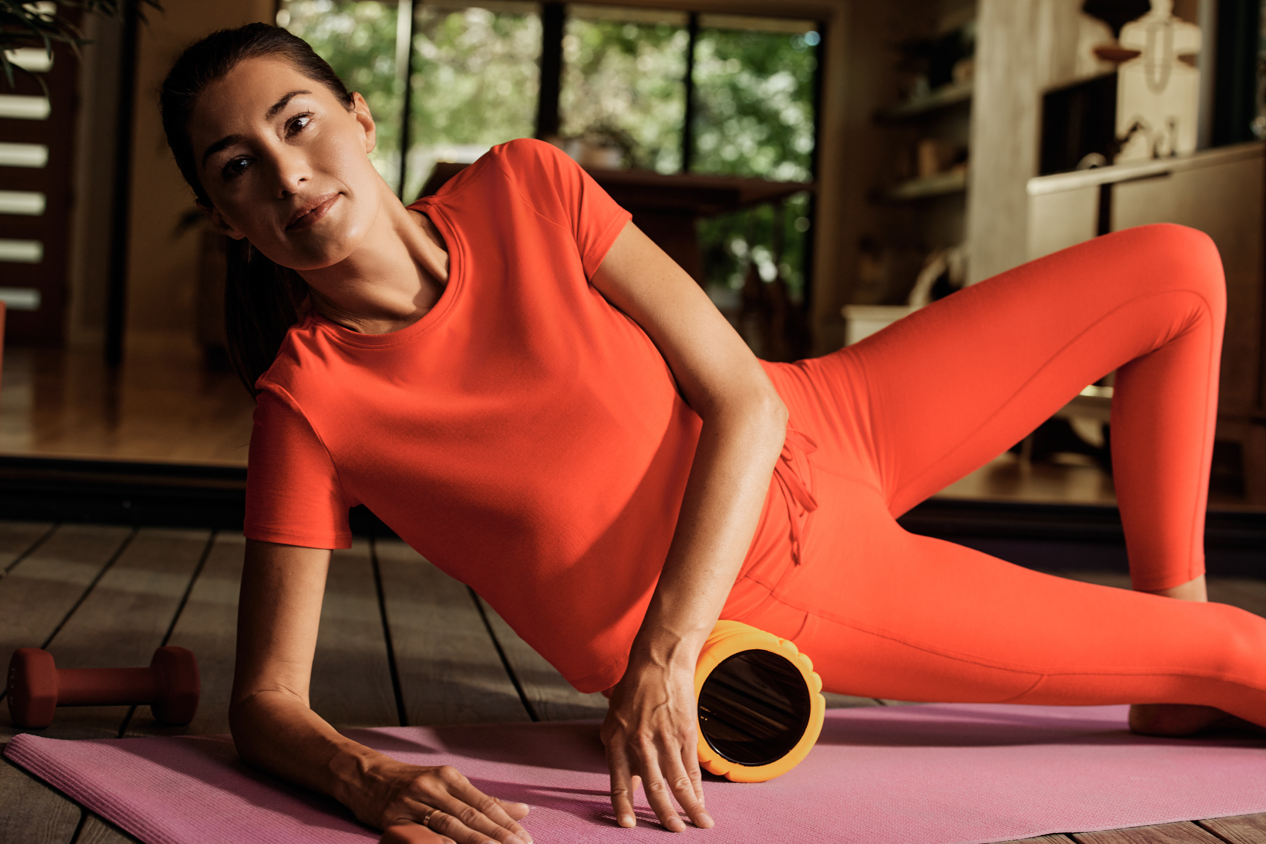 A woman in a bright orange athletic set performs a side stretch on a pink yoga mat, using a yellow foam roller. A dumbbell is visible on the wooden floor in the background of a sunlit home gym.