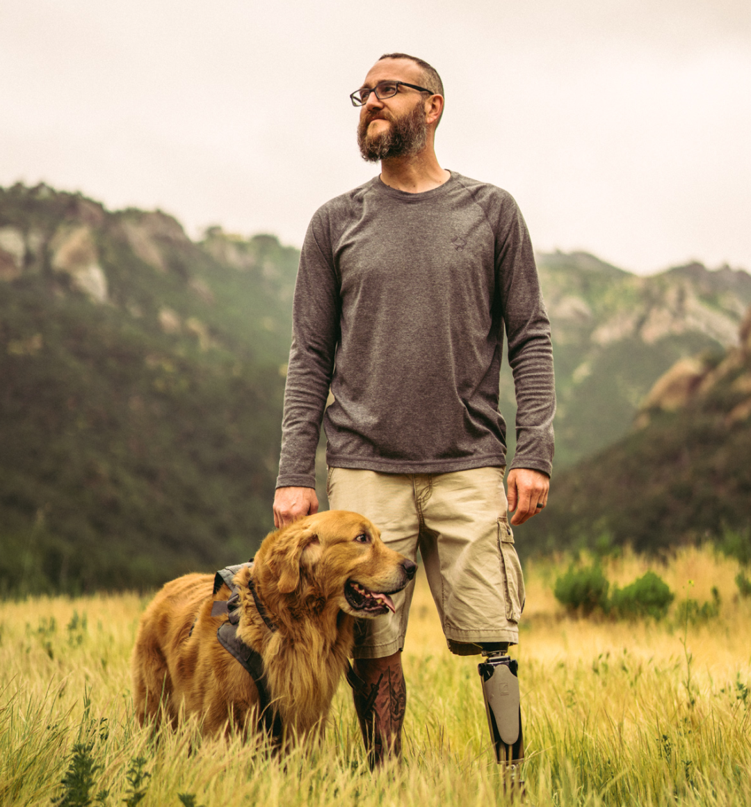 Man with a prosthetic leg standing in a field with a dog, mountains in the background