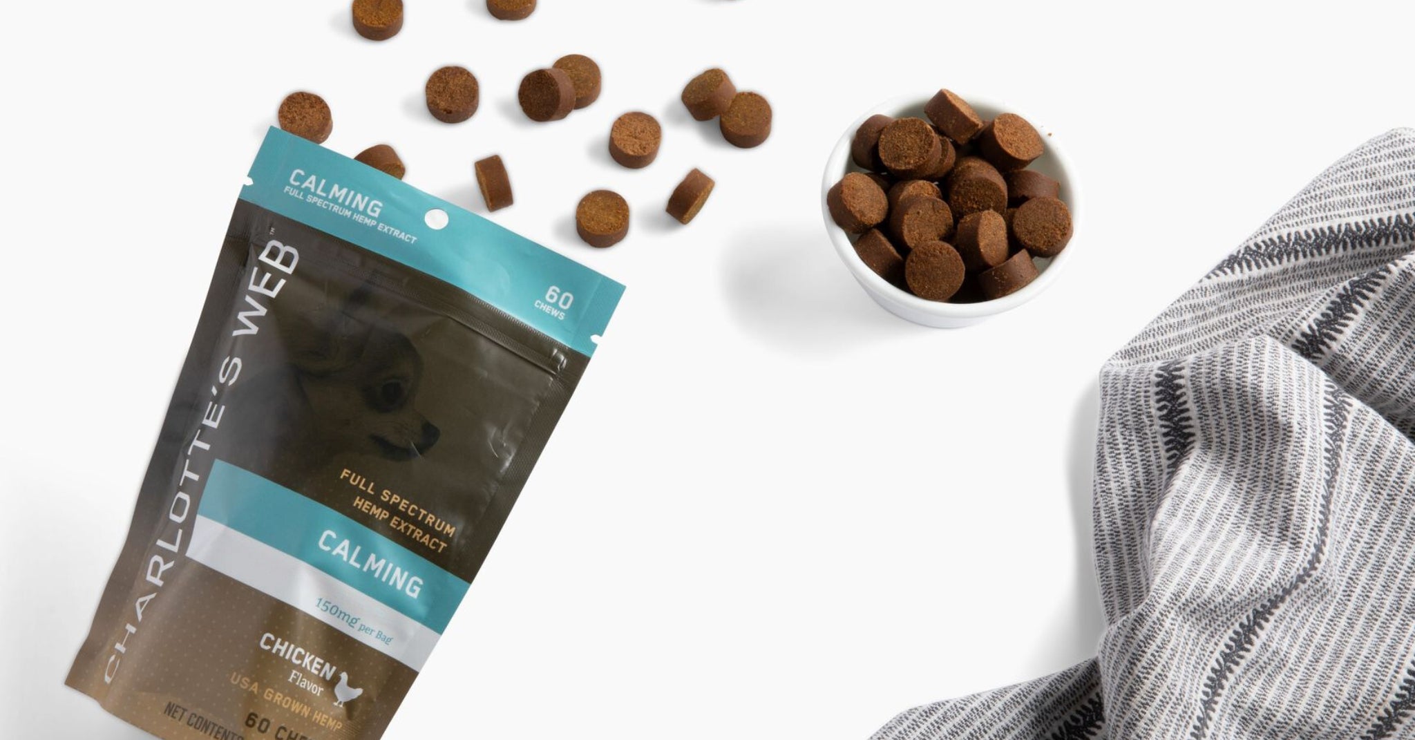 Charlotte's Web Calming Chicken-flavored hemp chews pouch partially opened beside a white bowl filled with chews, with extras scattered on a cozy patterned fabric background.