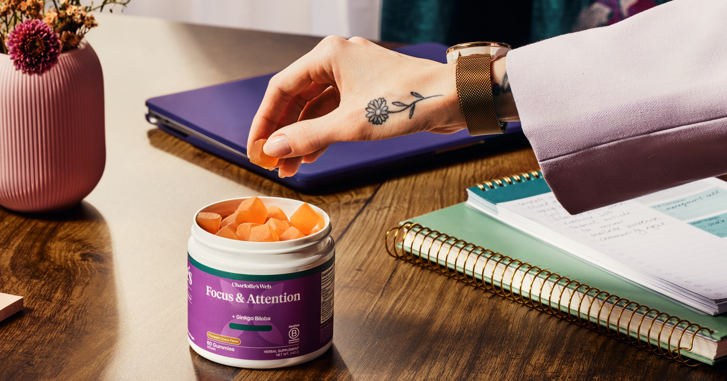 Person taking a supplement from a container labeled 'Focus & Attention' on a desk with notebooks and a notebook.