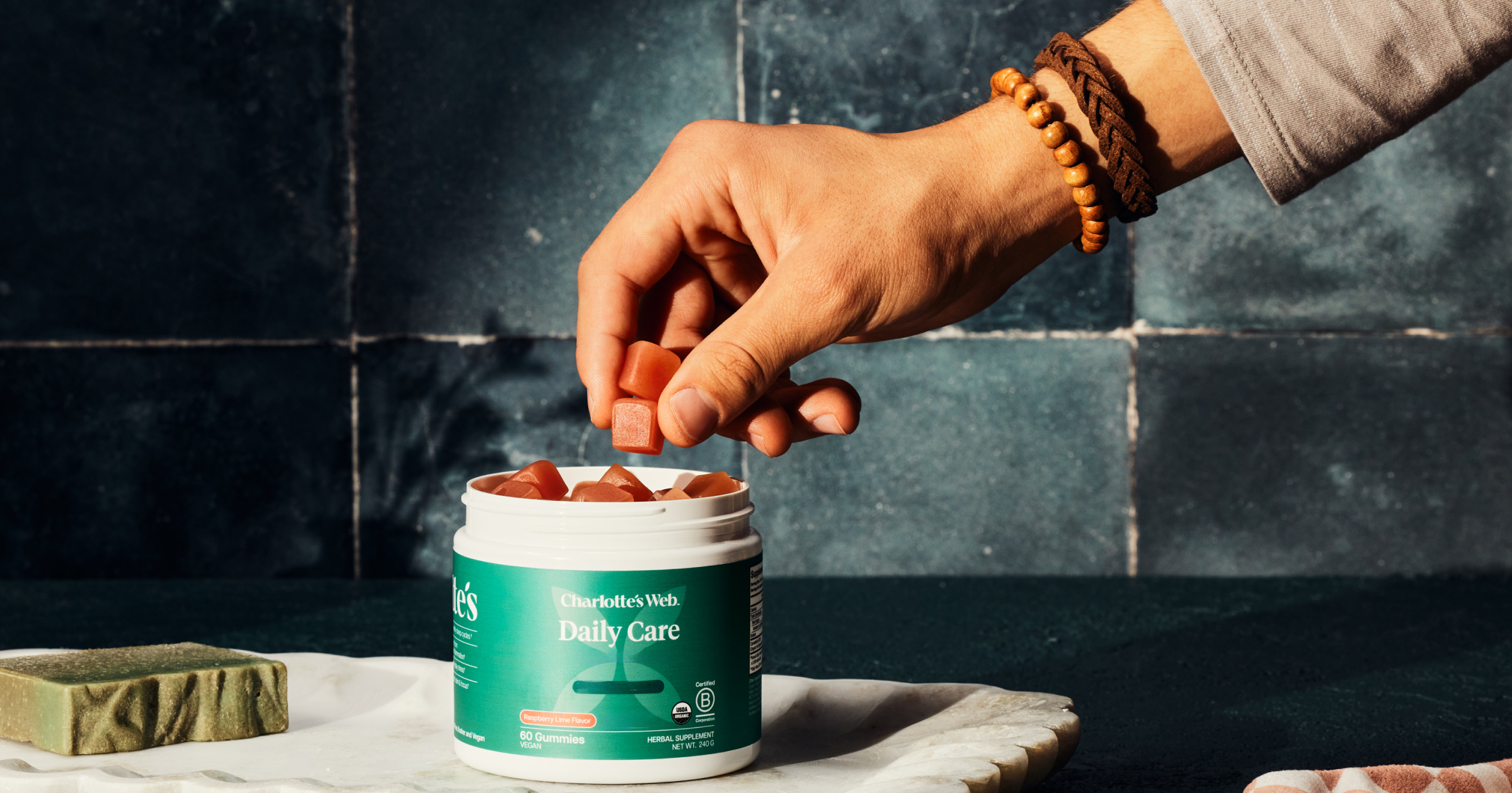 Person taking a supplement from a container labeled 'Daily Care' on a dark tiled wall background.