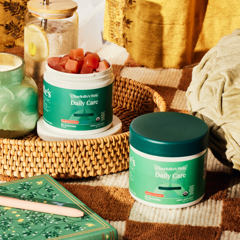Two containers of Charlotte's Web Daily Care on a textured surface with a woven basket and books in the background.