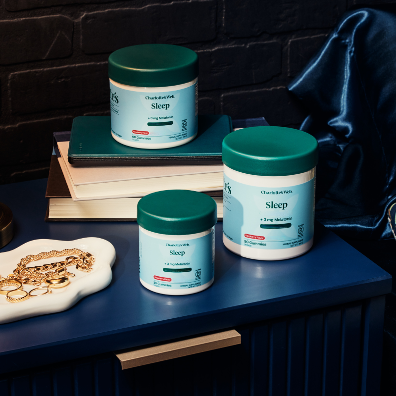 Three containers of Charlotte's Web Sleep Gummies on a blue table with books and jewelry.