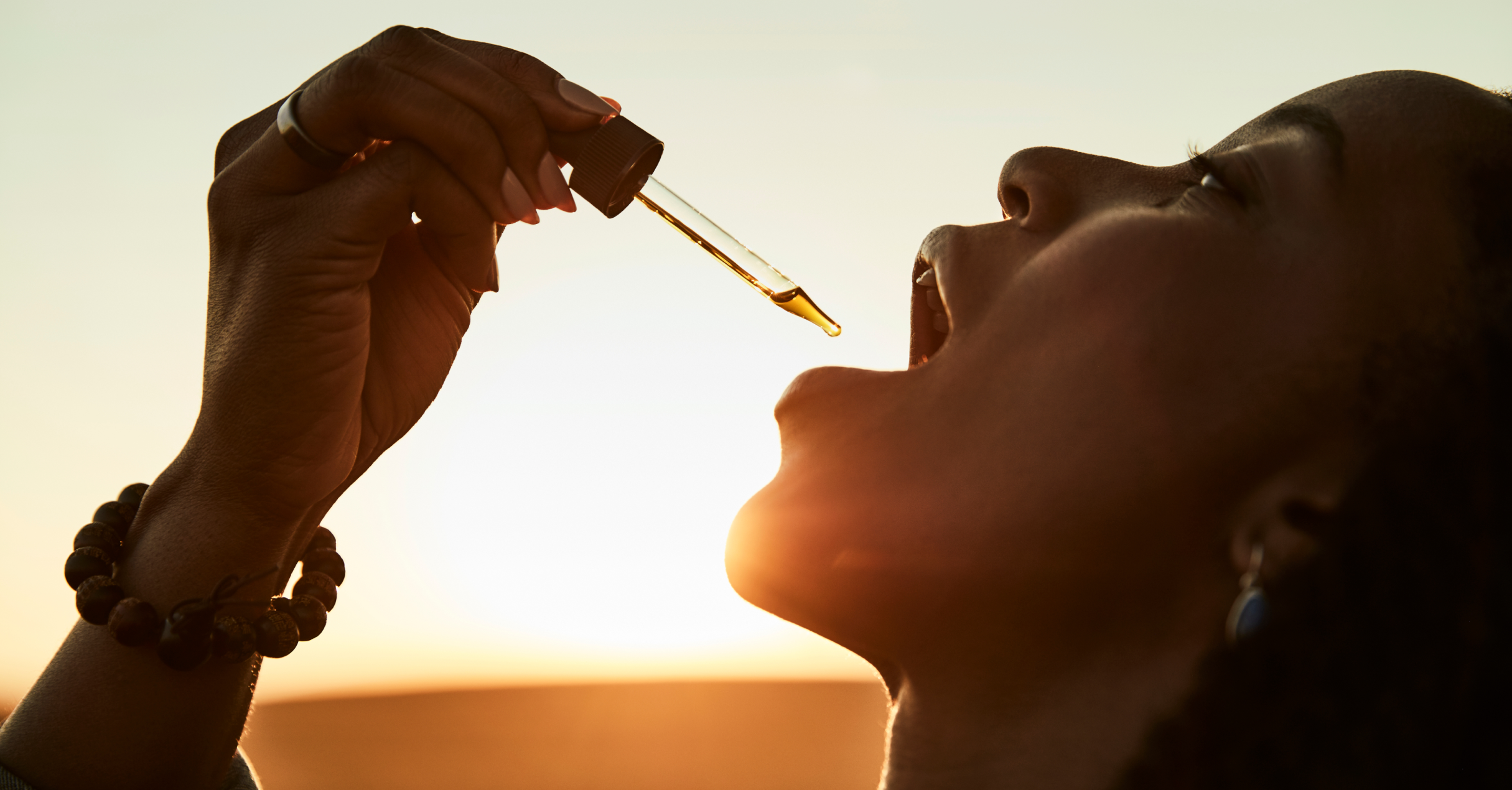 Person applying a dropper of liquid to their mouth against a sunset background