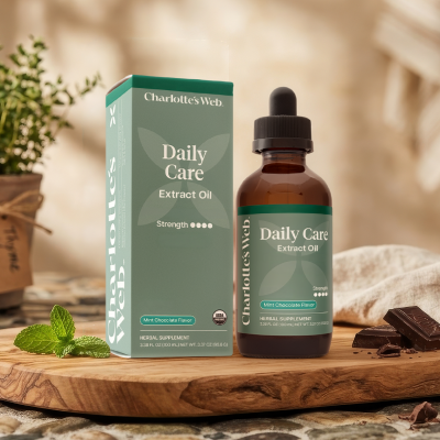 Charlotte's Web Daily Care Extract Oil bottle and matching box displayed on a wooden board with mint leaves and chocolate pieces, in a warm, softly blurred indoor setting.