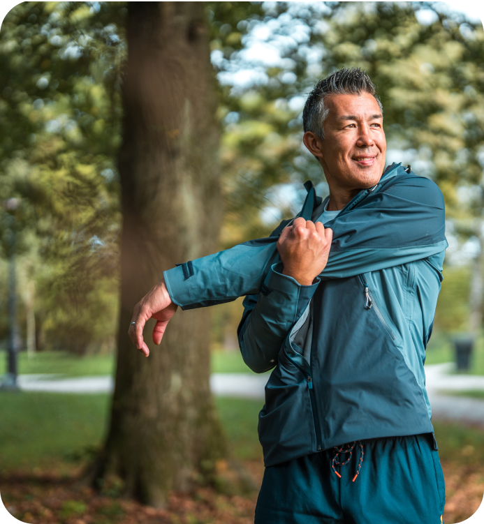 A smiling man outdoors in a park, stretching his arm while wearing a teal athletic jacket and matching teal sweatpants.