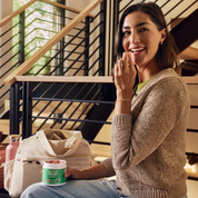 Product Image for Woman sitting on a staircase holding a container of yogurt, with a pink water bottle and beige bag nearby.