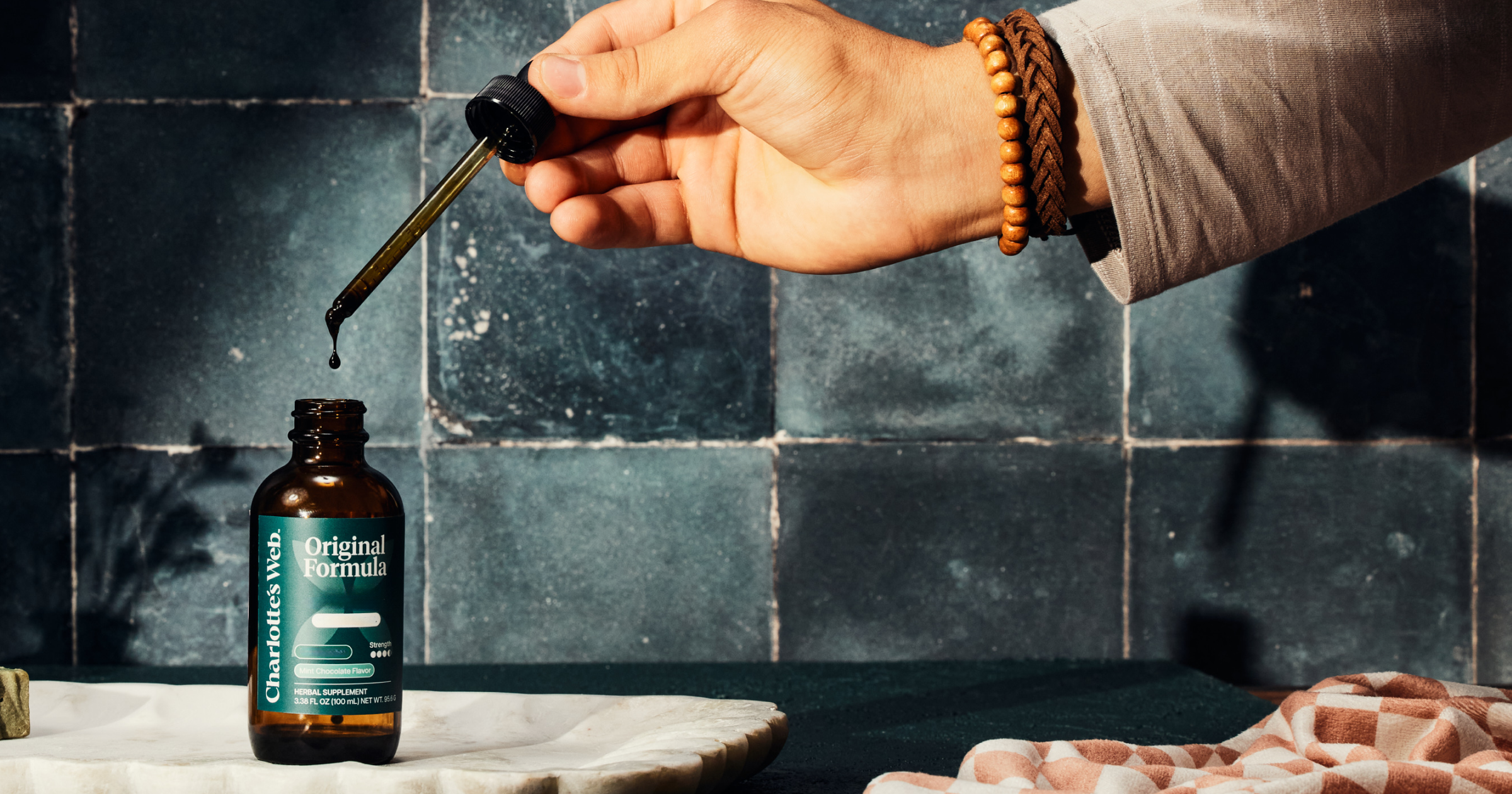 Hand pouring liquid from a dropper into a bottle labeled 'Original Formula' against a dark tiled wall.