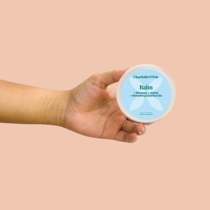 A person holding a container of Charlotte's Web Balm against a solid peach-colored background.