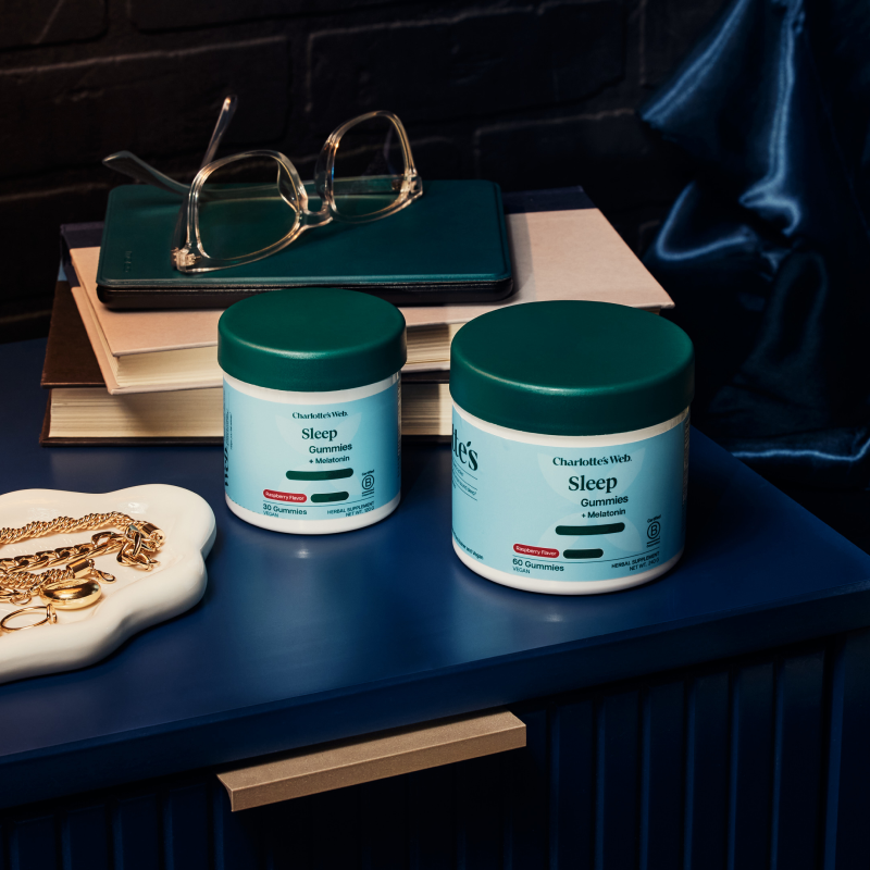 Two jars of Charlotte's Web Sleep Gummies displayed on a dark blue wooden table alongside books, eyeglasses, and a gold necklace, set against a dark brick wall and blue satin fabric.