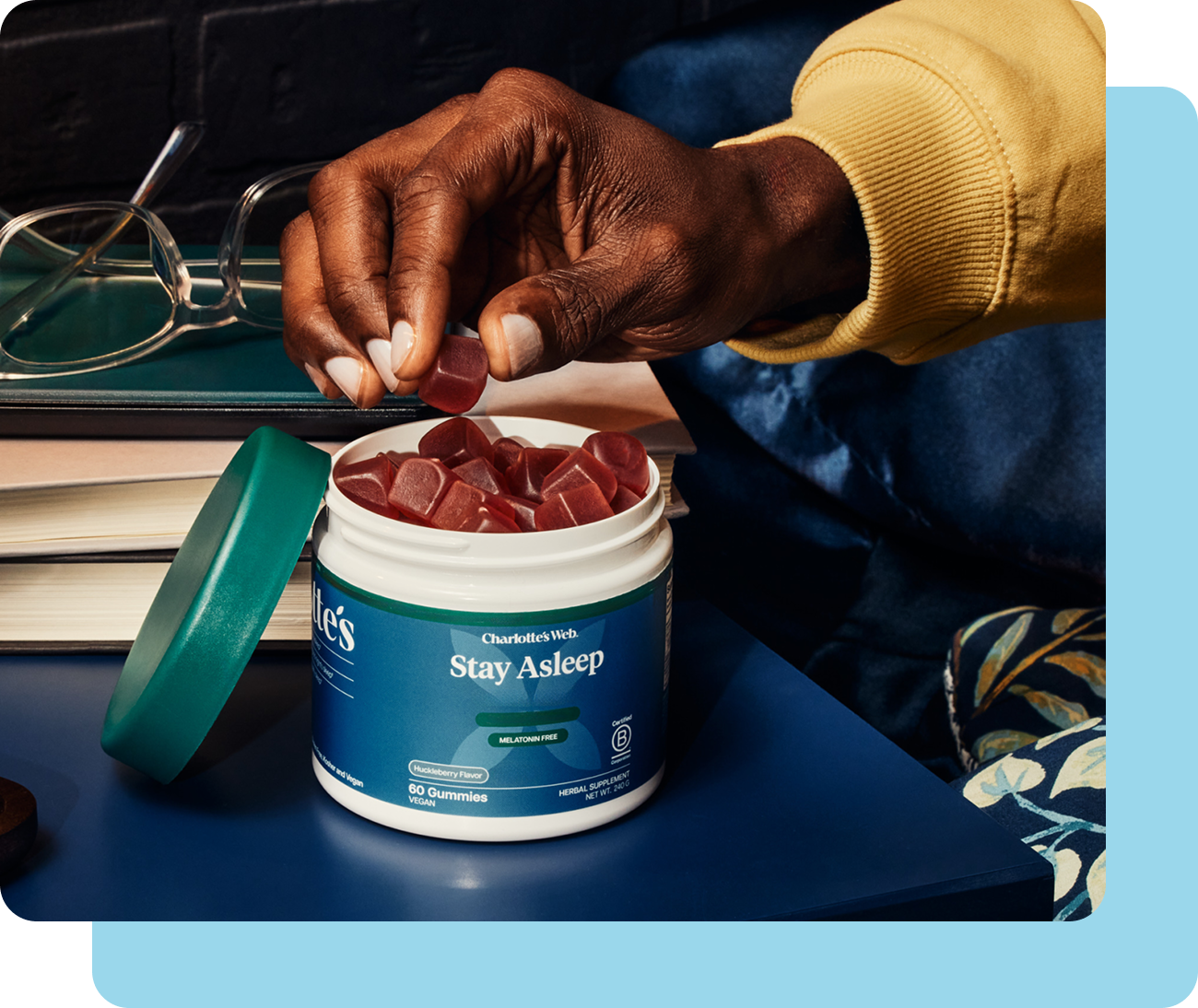 Person taking a supplement from a container labeled 'Stay Asleep' with glasses and books in the background.