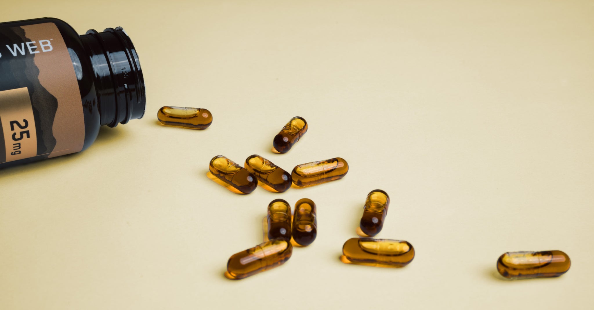 Black Charlotte's Web bottle labeled "WEB 25 mg" lying on its side with amber CBD softgel capsules spilled across a light yellow surface.
