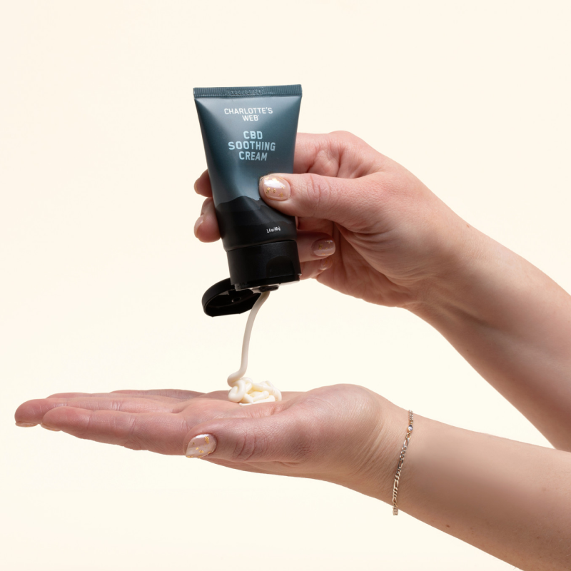 Hand holding a tube of Charlotte's Web CBD Soothing Cream with cream being dispensed onto another hand.