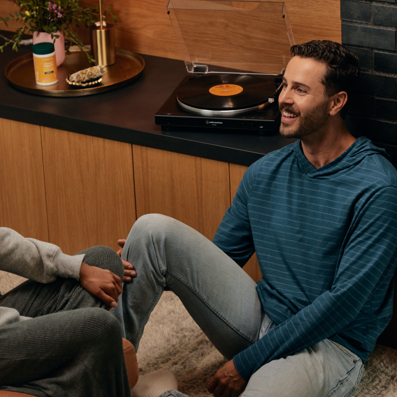 Man sitting on a rug in a cozy room with a record player and plants.