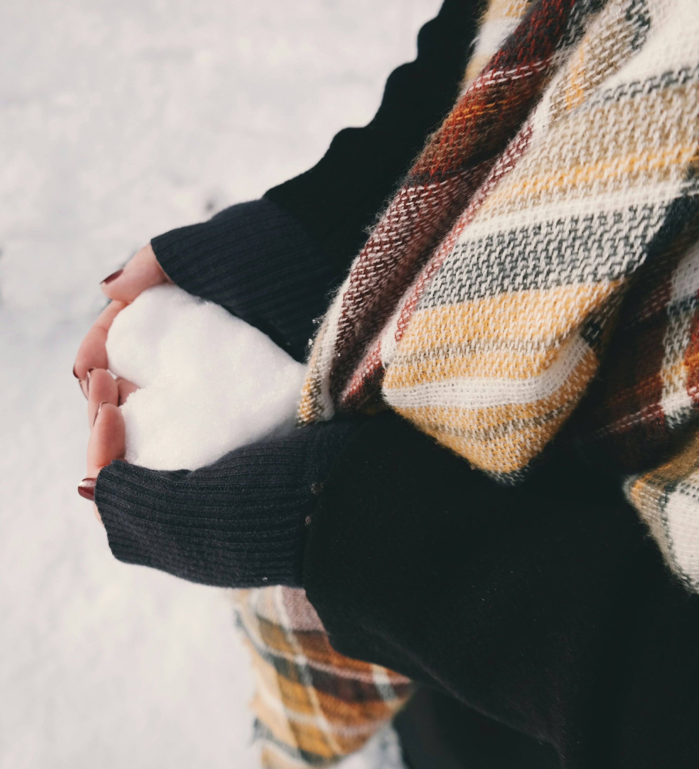 Person holding a snowball with both hands, dressed in a black sleeve and plaid scarf, set against a snowy winter background.