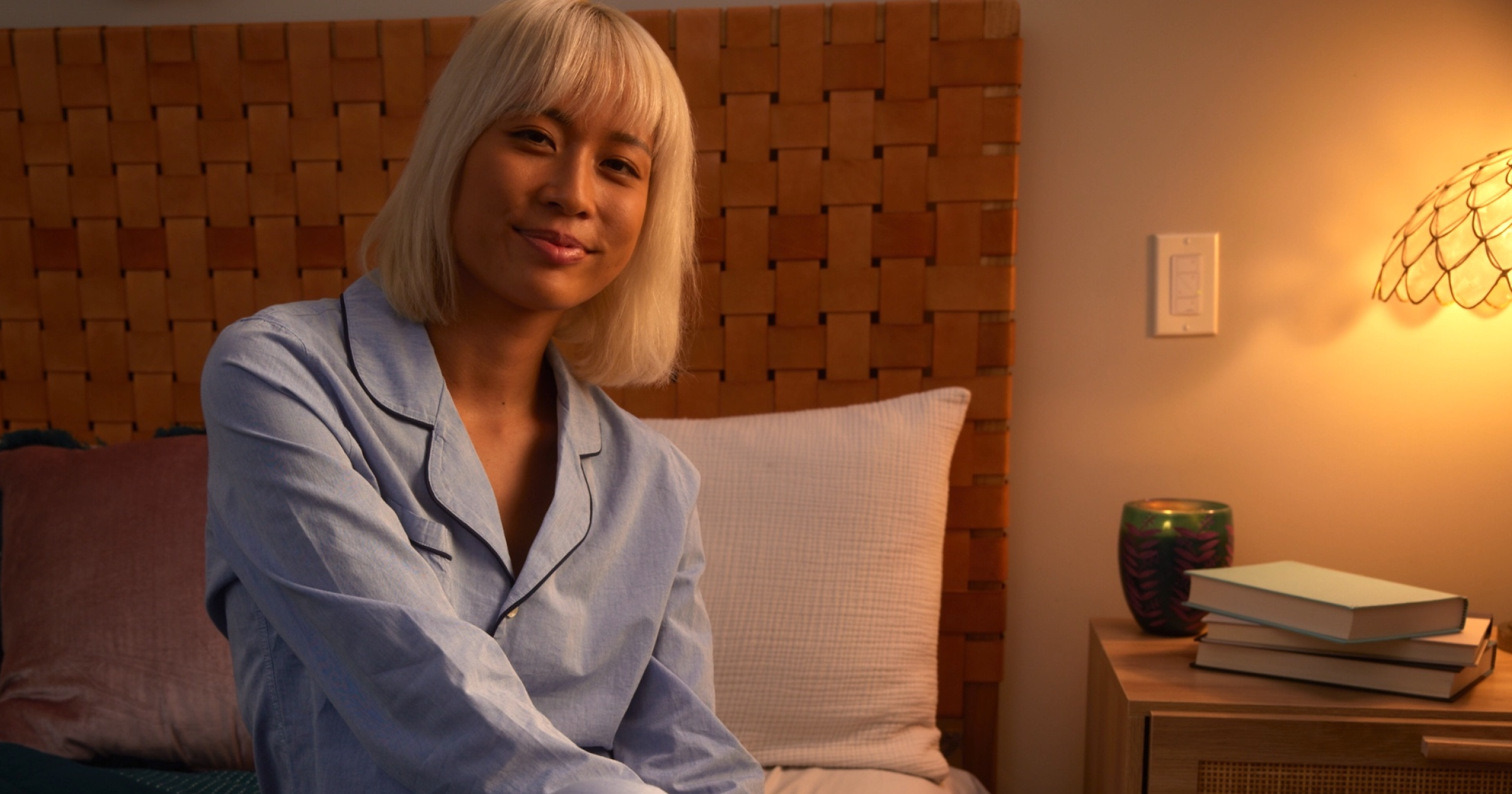 Woman in pajamas sitting on a bed in a cozy bedroom with warm lighting.