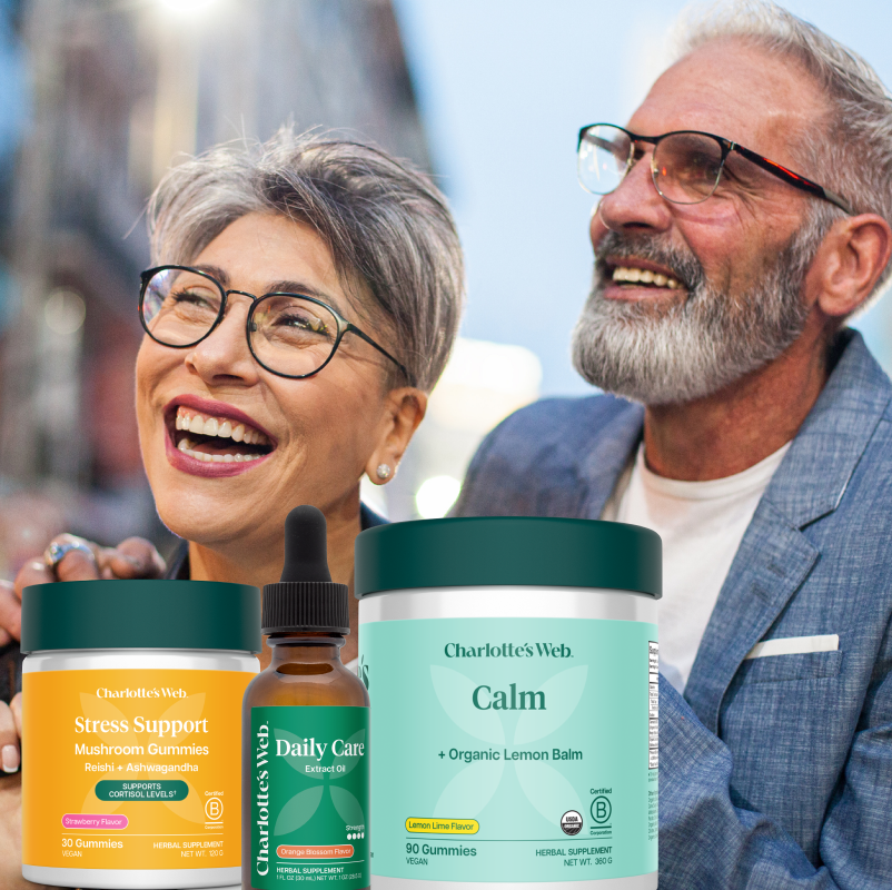 An elderly couple smiling and laughing outdoors, with three Charlotte's Web supplement containers in the foreground: Stress Support mushroom gummies in orange, Calm lemon balm gummies in mint green, and a small Daily Care tincture bottle.